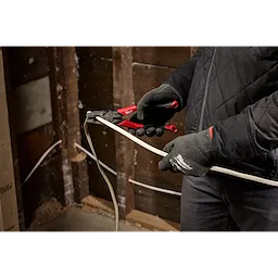 A person wearing gloves and a black jacket uses a pair of red-handled pliers to crimp an electrical wire inside a wooden stud wall under construction.