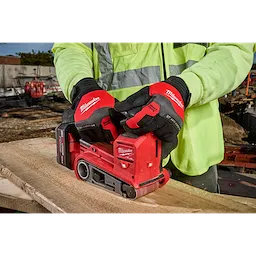 A person wearing Anti-Vibration Work Gloves operates a Milwaukee electric belt sander to smooth a wooden plank at a construction site. The gloves are red and black, designed to minimize vibration and improve grip during tool use. The person is also wearing a high-visibility yellow jacket.