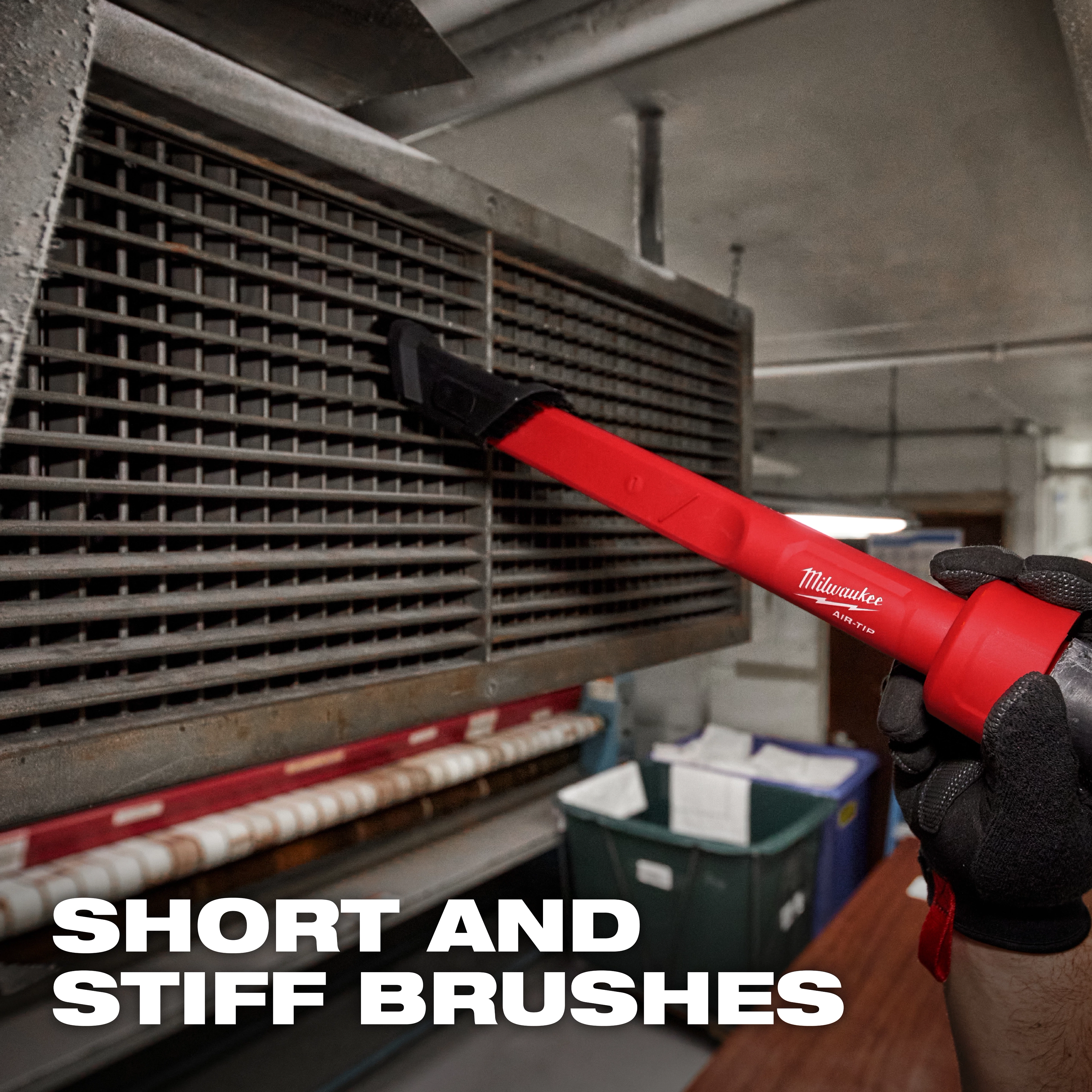 A person is using an AIR-TIP™ 3-in-1 Crevice and Brush Tool to clean the slats of a vent. The tool has a red handle and short, stiff brushes. The background includes industrial equipment and a caption reads "SHORT AND STIFF BRUSHES".