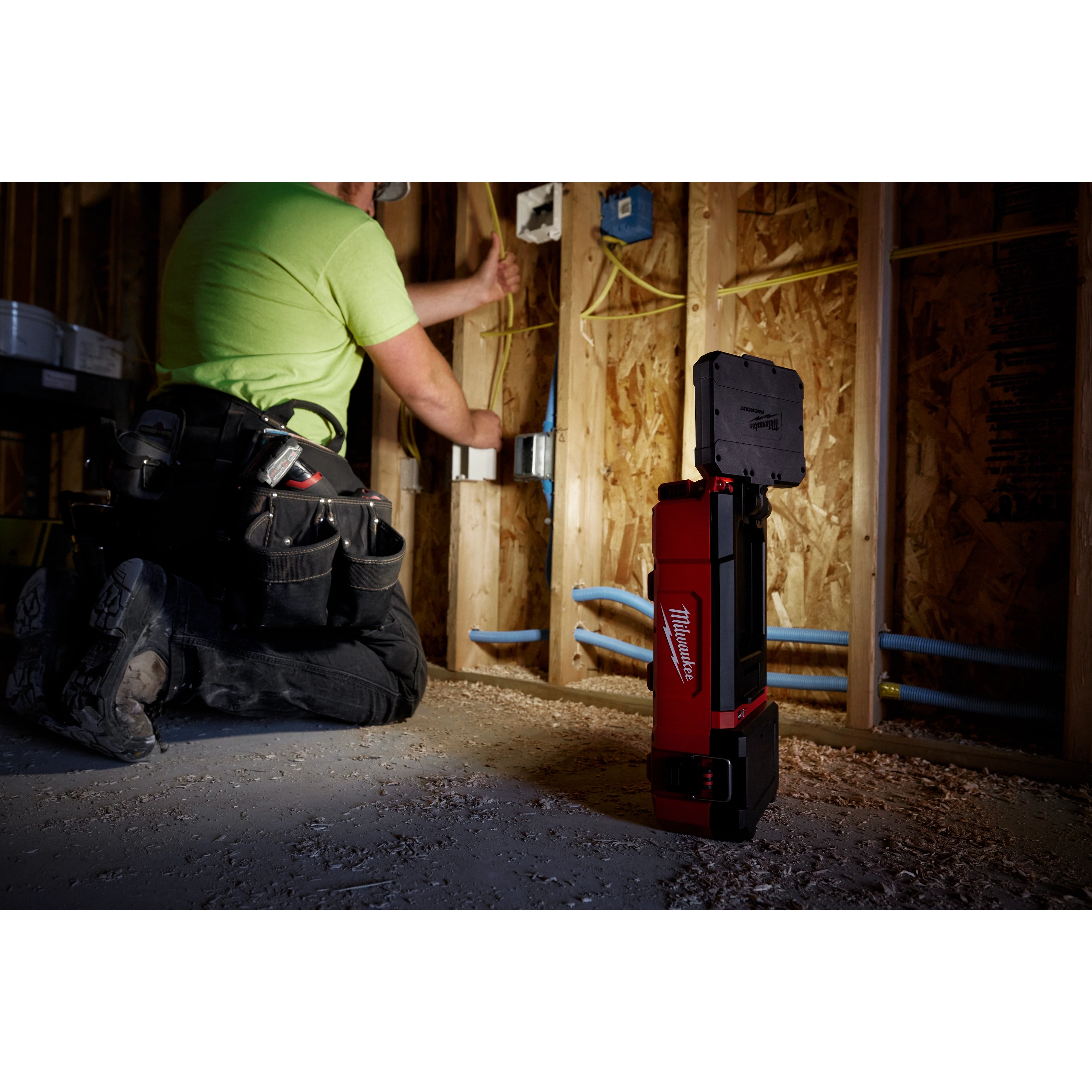 A worker uses the M12 PACKOUT Flood Light with USB Charging to illuminate a dimly lit construction area while installing wiring.
