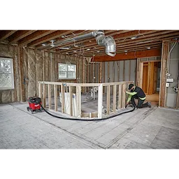 A worker using a 1-7/8” x 16' flexible hose connected to a red vacuum in a partially constructed home interior.