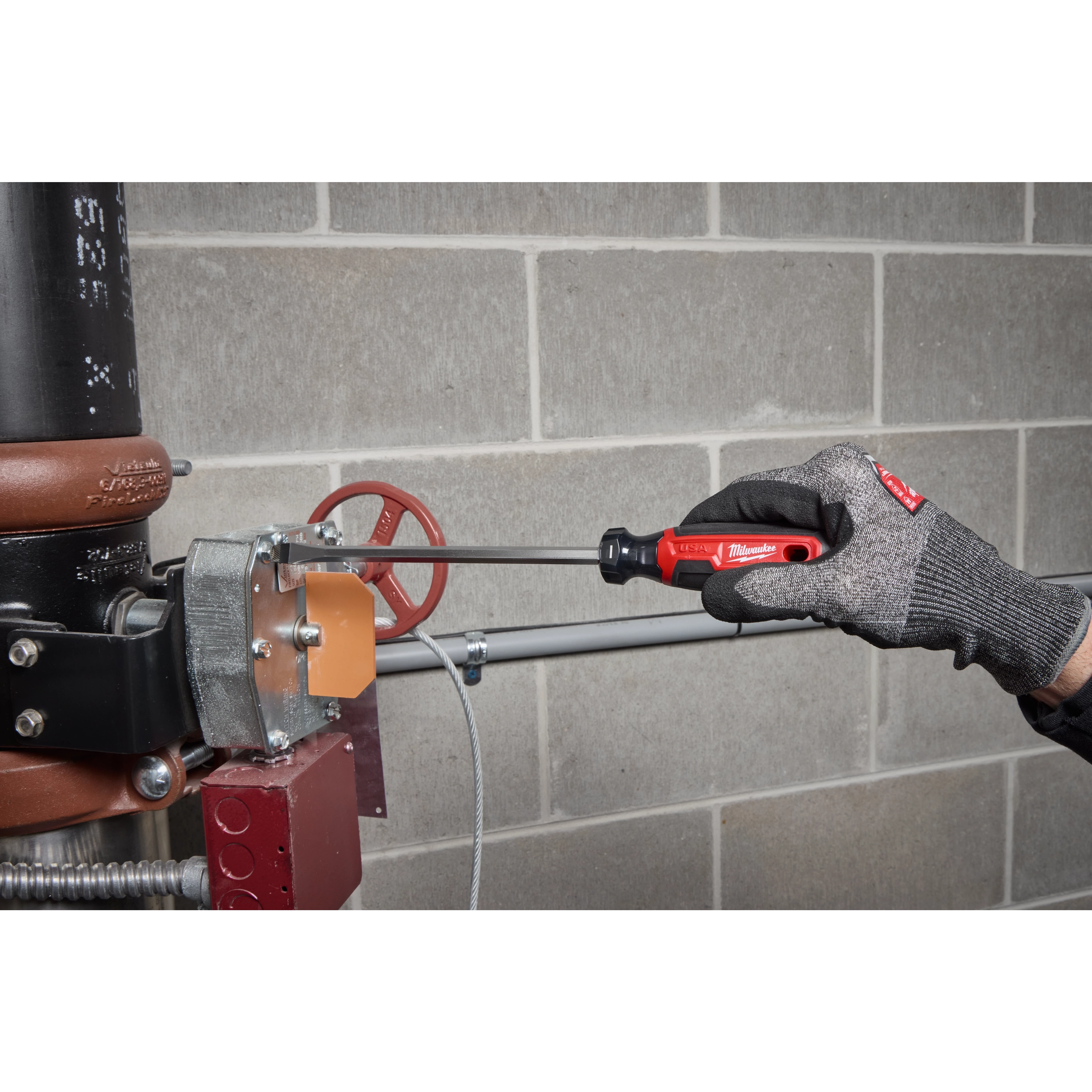 Gloved hand using a 3/8" Slotted 8" Cushion Grip Screwdriver (USA) on metal equipment against a block wall.