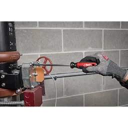 Gloved hand using a 3/8" Slotted 8" Cushion Grip Screwdriver (USA) on metal equipment against a block wall.
