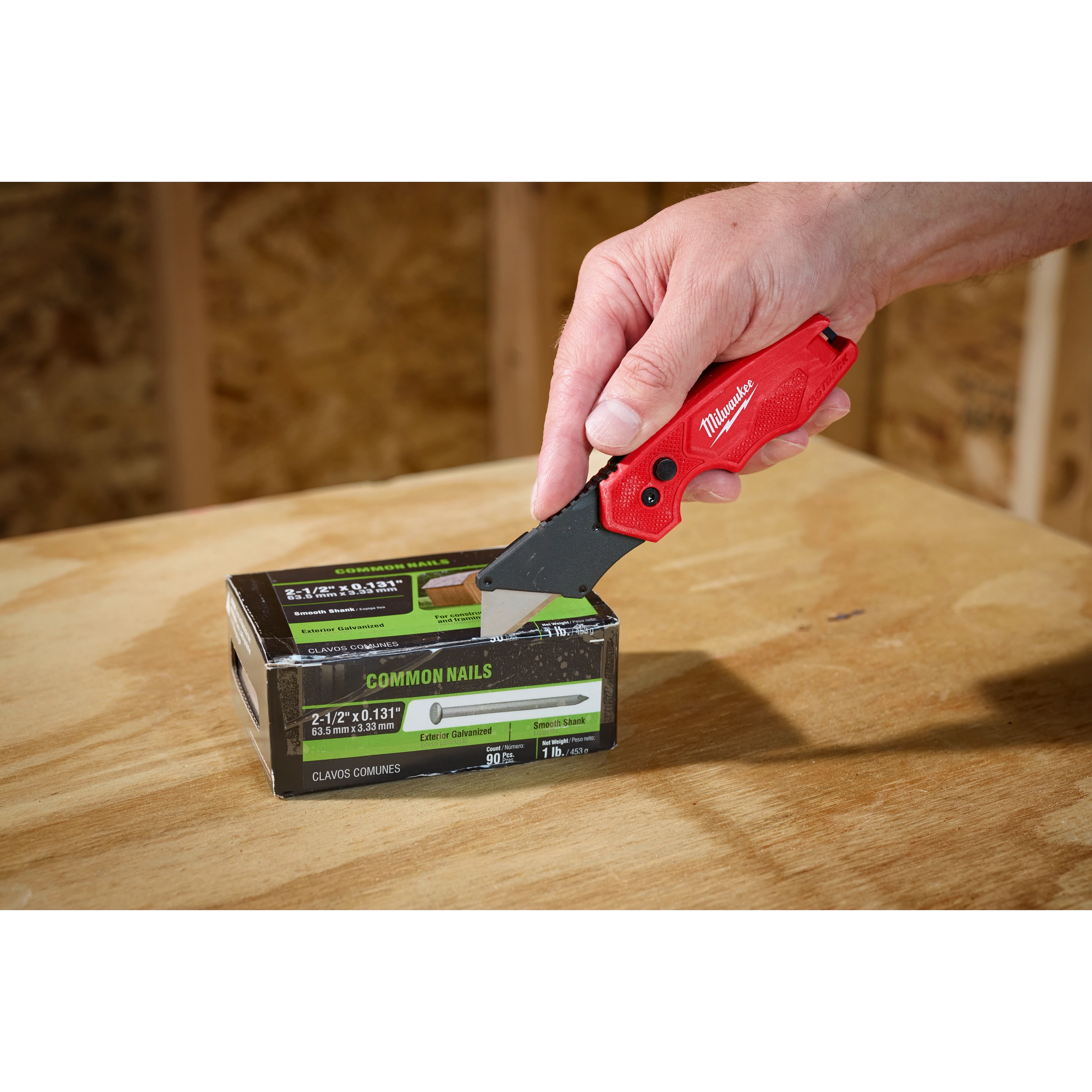 Hand using a FASTBACK Compact Folding Utility Knife to cut into a box of common nails on a wooden surface.
