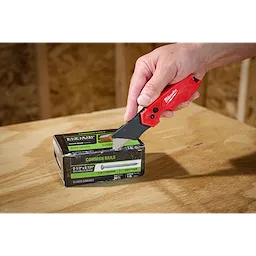 Hand using a FASTBACK Compact Folding Utility Knife to cut into a box of common nails on a wooden surface.