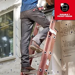 A man wearing gray Work Pants with a durable 7-pocket design is climbing a red ladder outside a building under construction. The pants have multiple pockets and tools attached, highlighting their practicality for work environments.