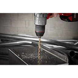 The RED HELIX™ Cobalt 3/8" Drill Bit is attached to a drill and is actively drilling into a metal surface, creating fine metal shavings. The drill is held by a hand and positioned above a square metal object, with industrial pipes in the background.