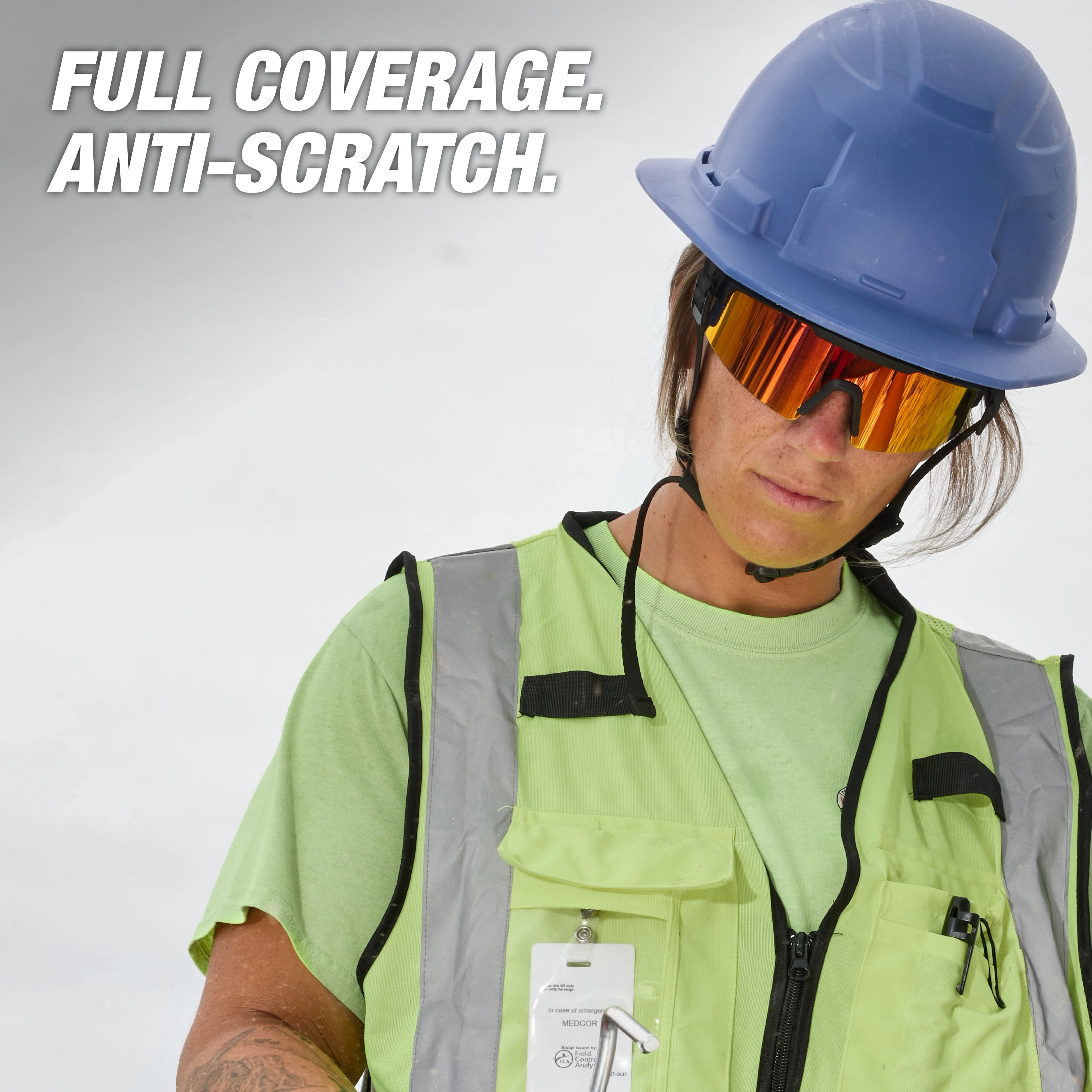 Worker wearing wrap-around safety glasses, a green safety vest with reflective strips, and a blue hard hat. The image has the text "Full Coverage. Anti-Scratch."