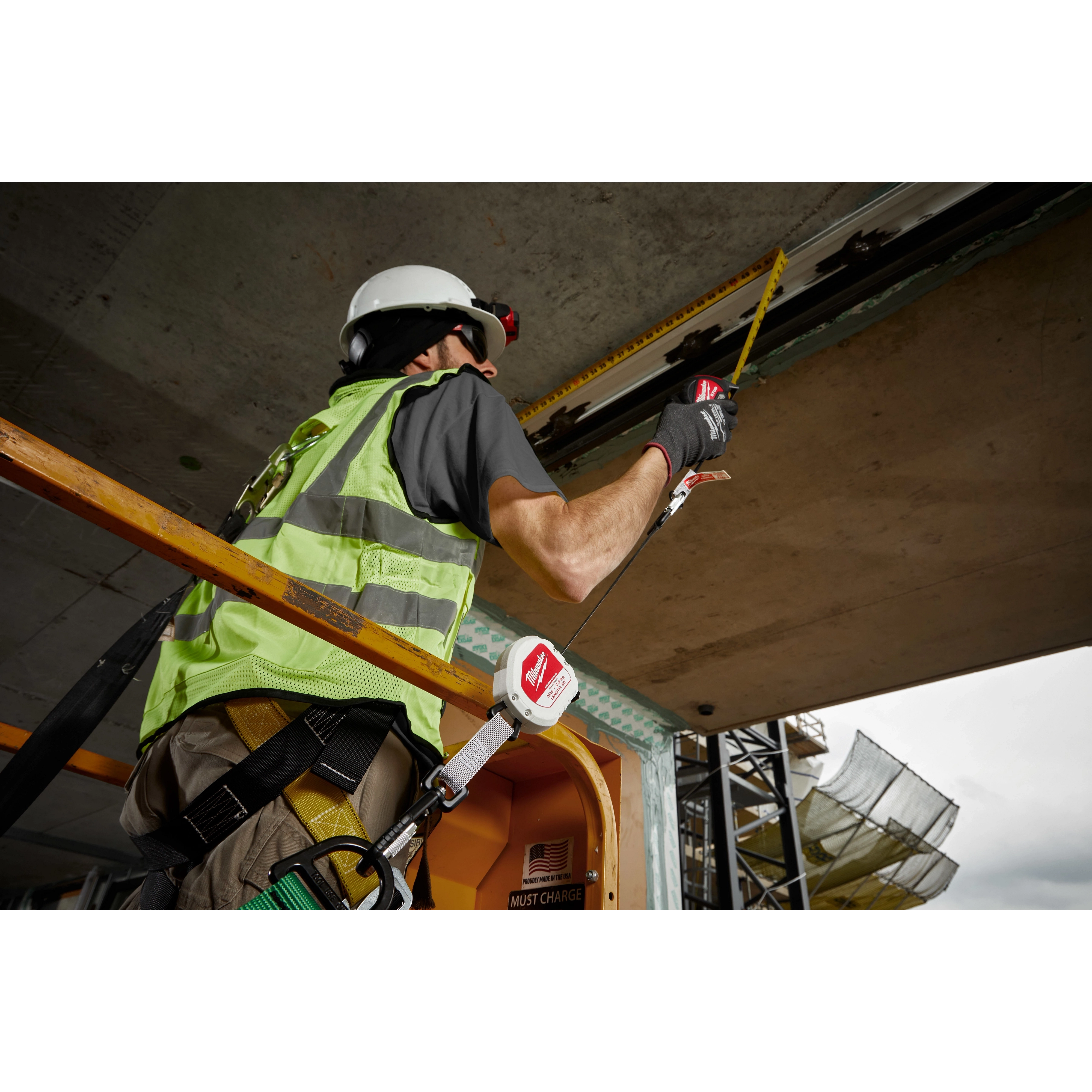 A construction worker using a tape measure while standing on a lift, secured with a safety harness. Attached to the worker's harness is a 5LB Quick-Connect Retractable Tool Lanyard, featuring a black and green cord and red logo. The worker wears protective gloves, a helmet, and a safety vest.
