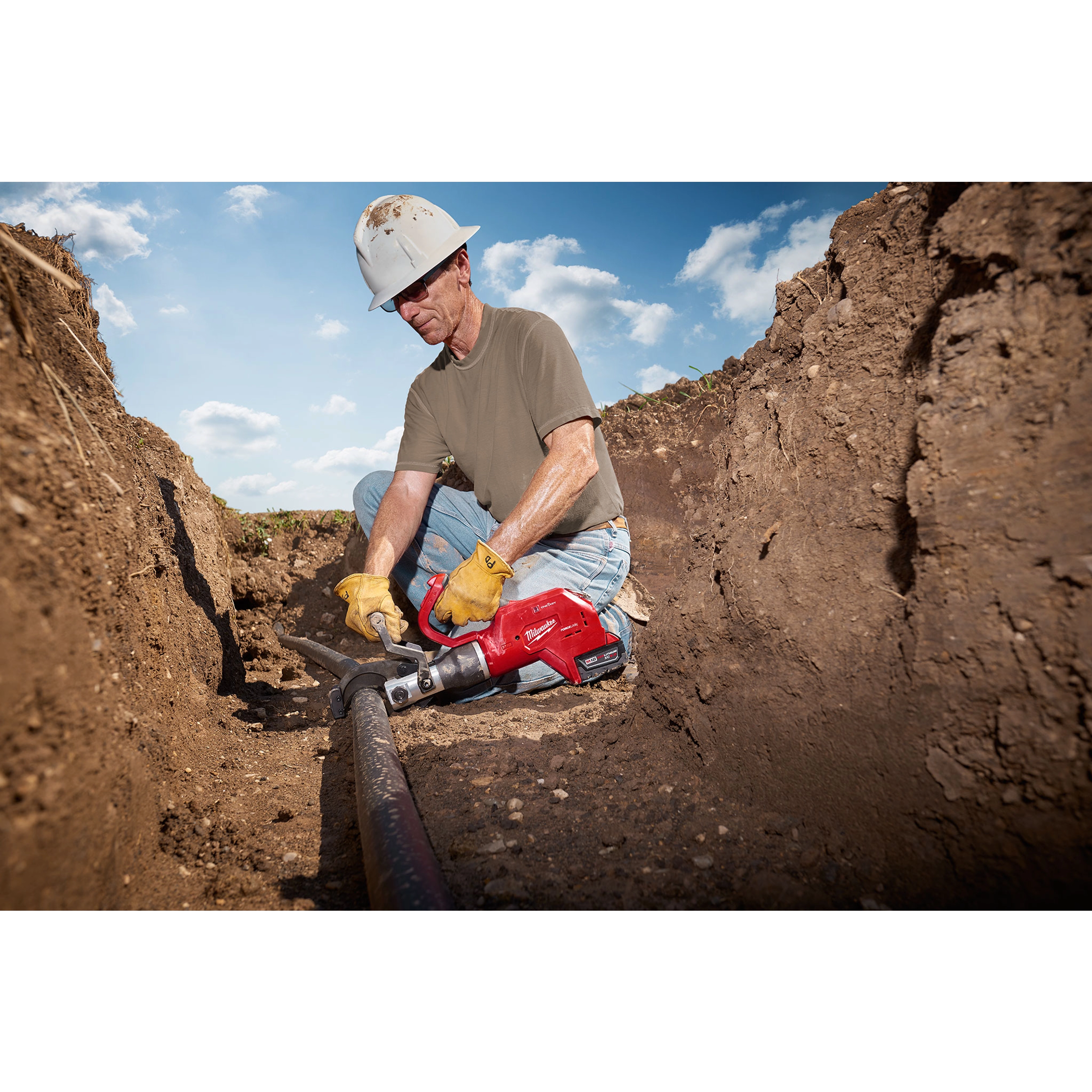 A worker uses the FORCELOGIC™ M18™ 3" Underground Cable Cutter Kit to cut a large underground cable. The tool is red and designed for heavy-duty industrial use. The worker is wearing yellow gloves, a white hard hat, and sits in a trench on a sunny day.