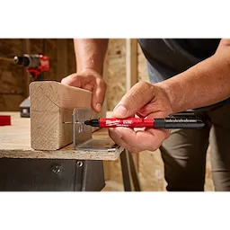 A person uses an INKZALL™ 2IN1 Deep Reach Ultra Fine Point Black Jobsite Marker to mark a metal bracket attached to a wooden beam. The red marker has a pocket clip and is held with one hand, with the other hand stabilizing the wood.