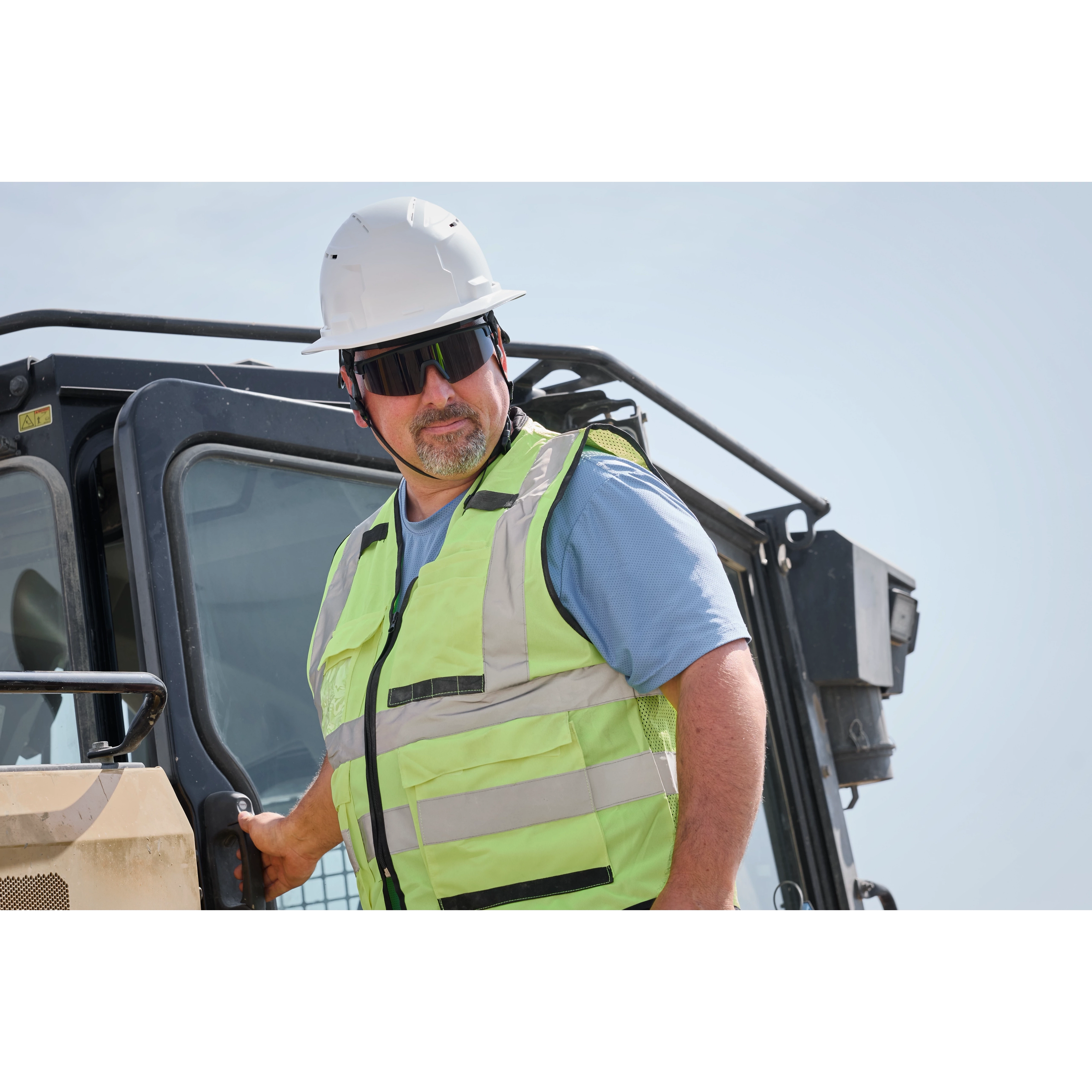 A construction worker in a high-visibility safety vest and white hard hat stands next to heavy machinery. The person is wearing Wrap Around Safety Glasses with Tinted Anti-Scratch Lenses.