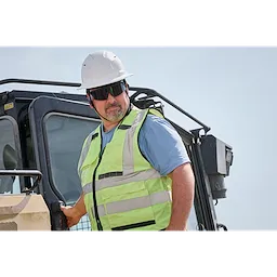 A construction worker in a high-visibility safety vest and white hard hat stands next to heavy machinery. The person is wearing Wrap Around Safety Glasses with Tinted Anti-Scratch Lenses.