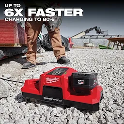 Milwaukee Supercharger is displayed on a construction site, emphasizing its ability to charge batteries up to six times faster to 80%. The bright red charger, equipped with a battery, stands on gravel near a worker and construction materials, highlighting its efficiency in demanding environments.