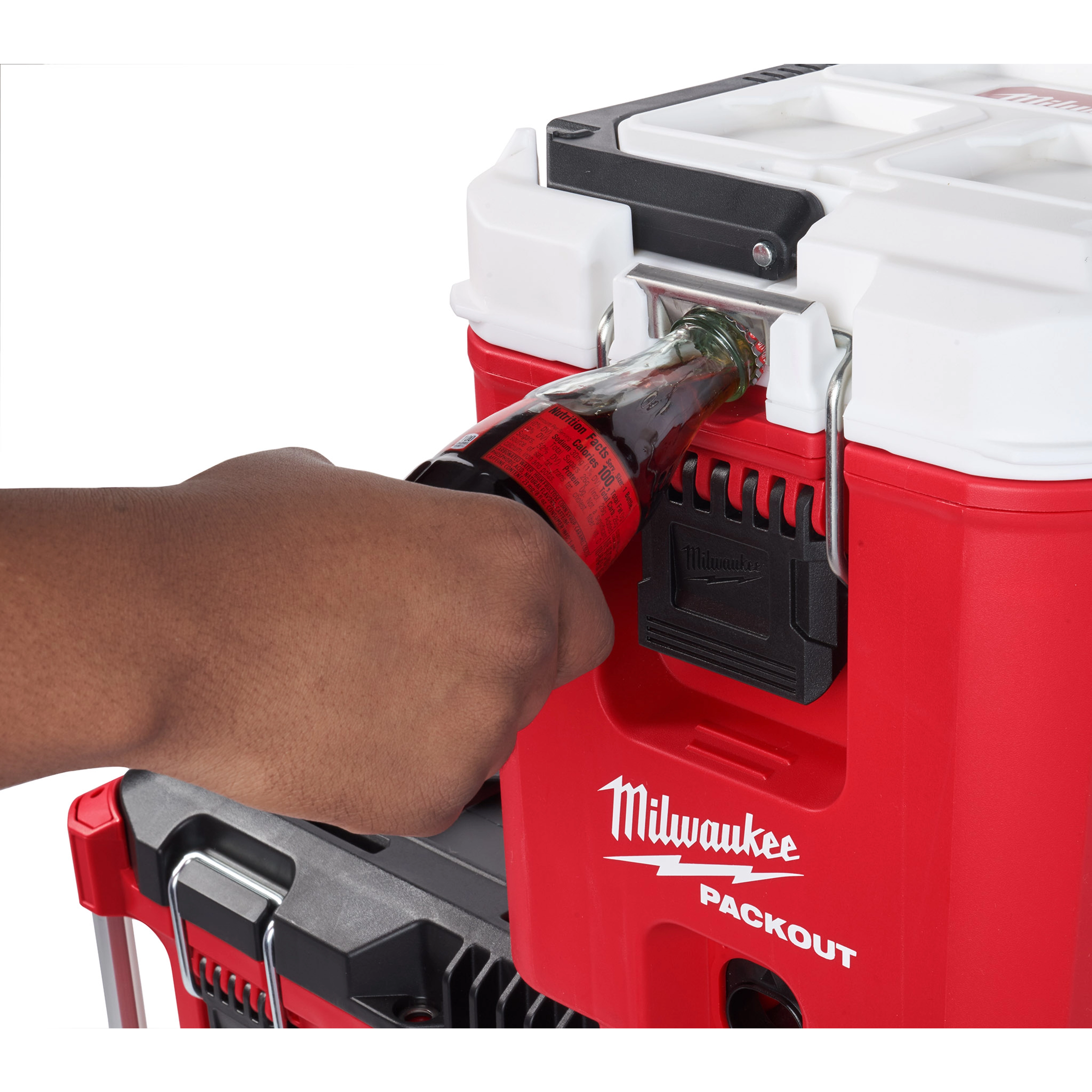 A person is using a built-in bottle opener on a red and white PACKOUT™ 16QT Compact Cooler. The cooler has the Milwaukee logo on the front and is stacked on another PACKOUT™ unit.