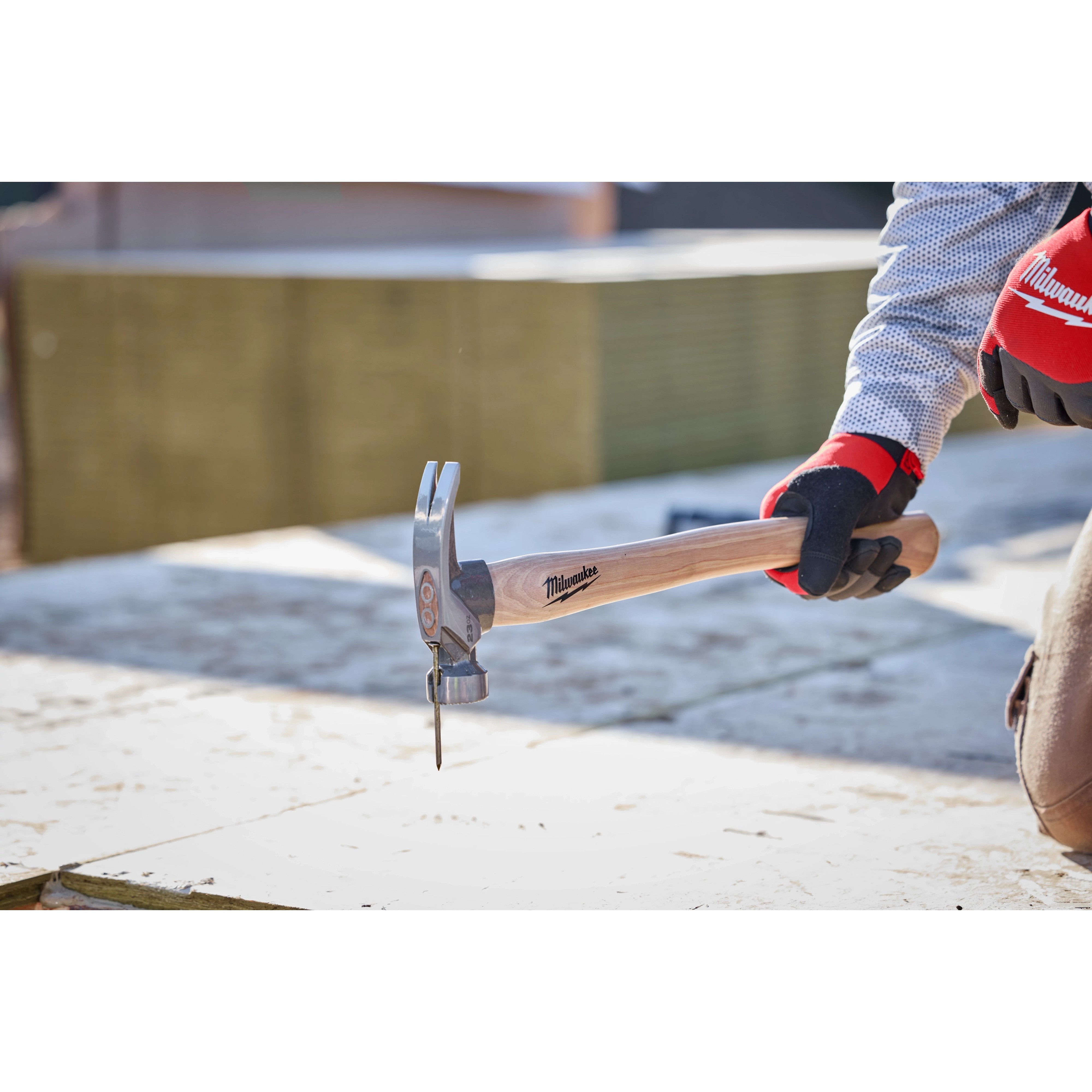 23oz Milled Face Hickory Handle Framing Hammer in use by a person wearing gloves, driving a nail into a wooden surface.