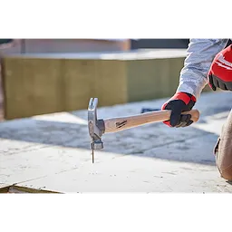 23oz Milled Face Hickory Handle Framing Hammer in use by a person wearing gloves, driving a nail into a wooden surface.