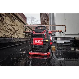 The PACKOUT™ Structured Backpack is shown in an industrial outdoor setting, resting on wet pavement. The black and red backpack features several compartments and the "Milwaukee" logo. It is designed for tools, with visible reinforcements and handles.