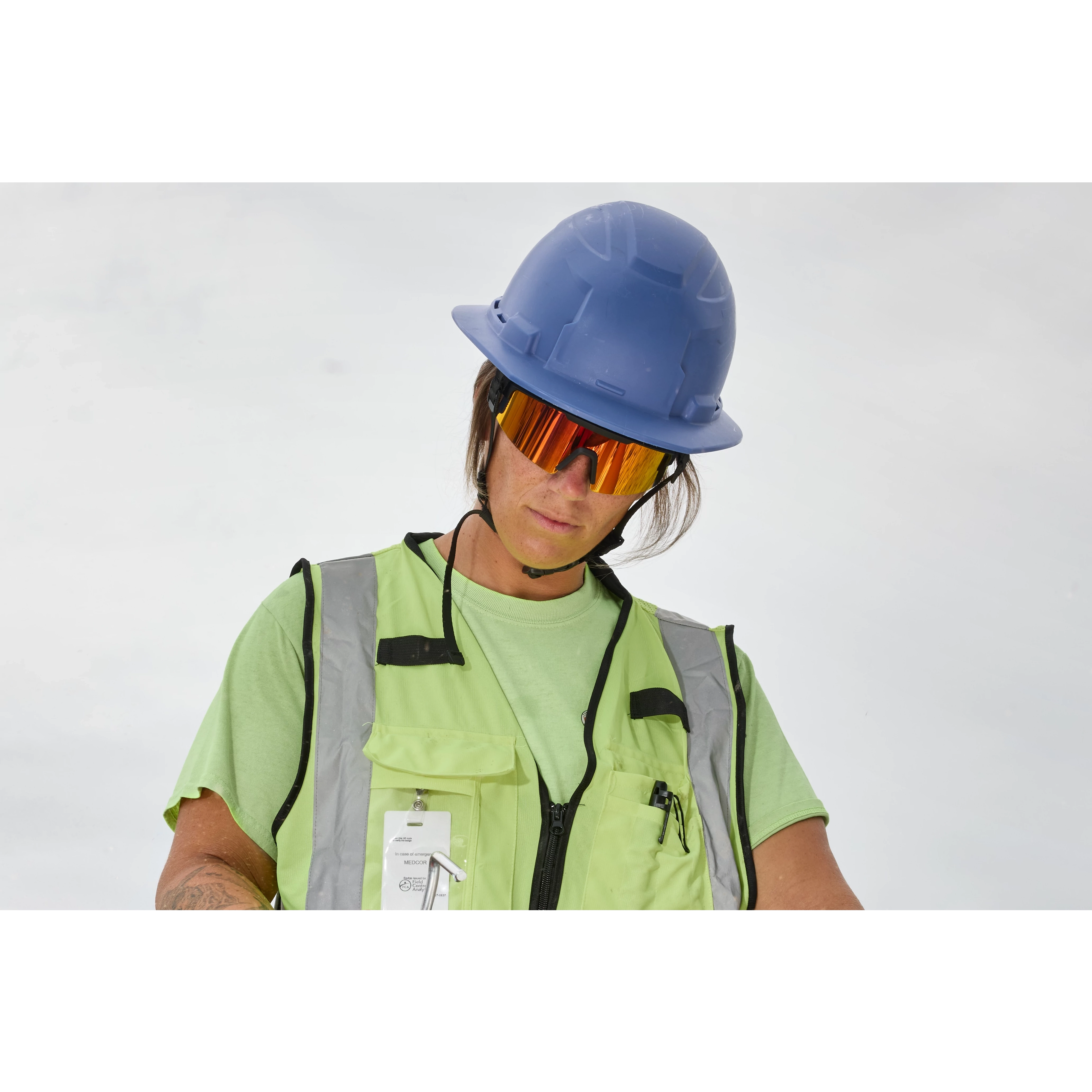 A person wearing a blue hard hat, light green safety vest with reflective strips, and holding a pair of Wrap Around Safety Glasses - Red Mirrored Anti-Scratch Lenses.