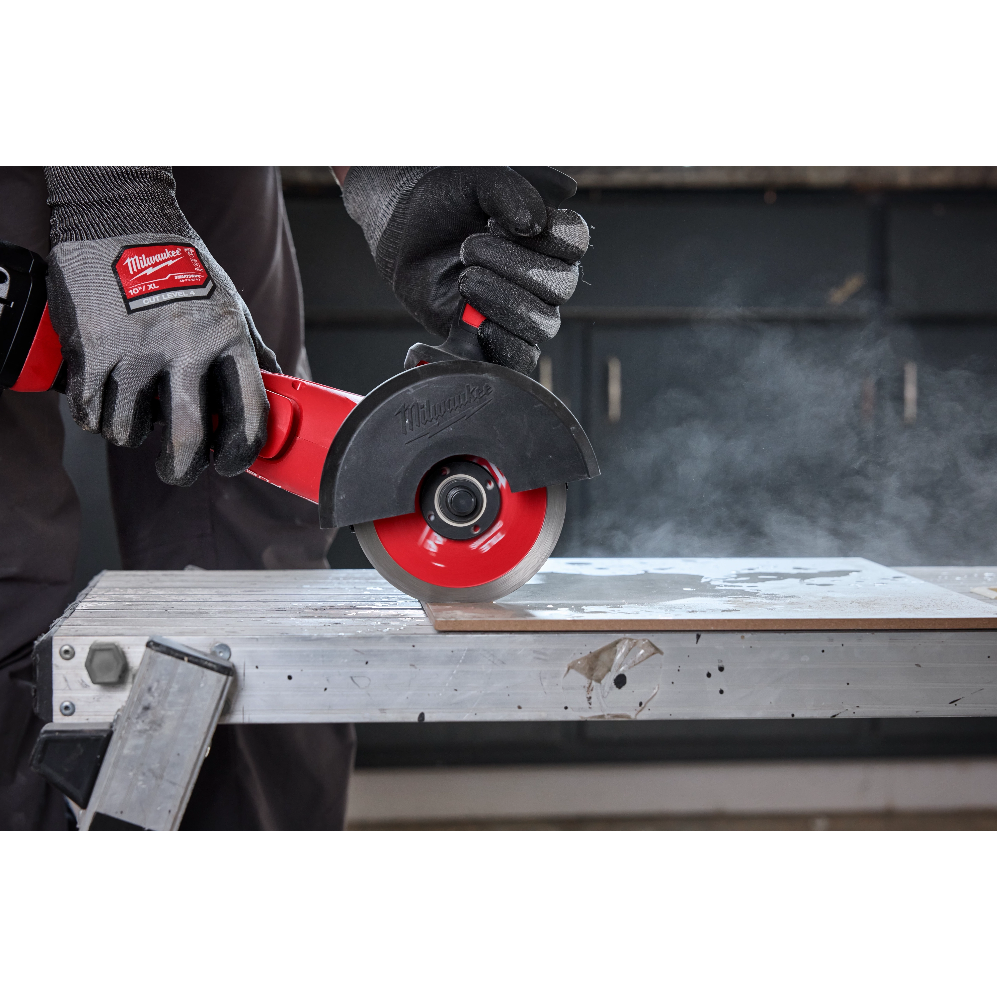 A gloved hand is using a Milwaukee tool equipped with a 4-1/2" Continuous Tile Diamond Blade to cut through a tile on a workbench. Dust is visible in the air from the cutting process. The blade is red and black, and the setting appears to be a workshop.