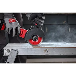 A gloved hand is using a Milwaukee tool equipped with a 4-1/2" Continuous Tile Diamond Blade to cut through a tile on a workbench. Dust is visible in the air from the cutting process. The blade is red and black, and the setting appears to be a workshop.