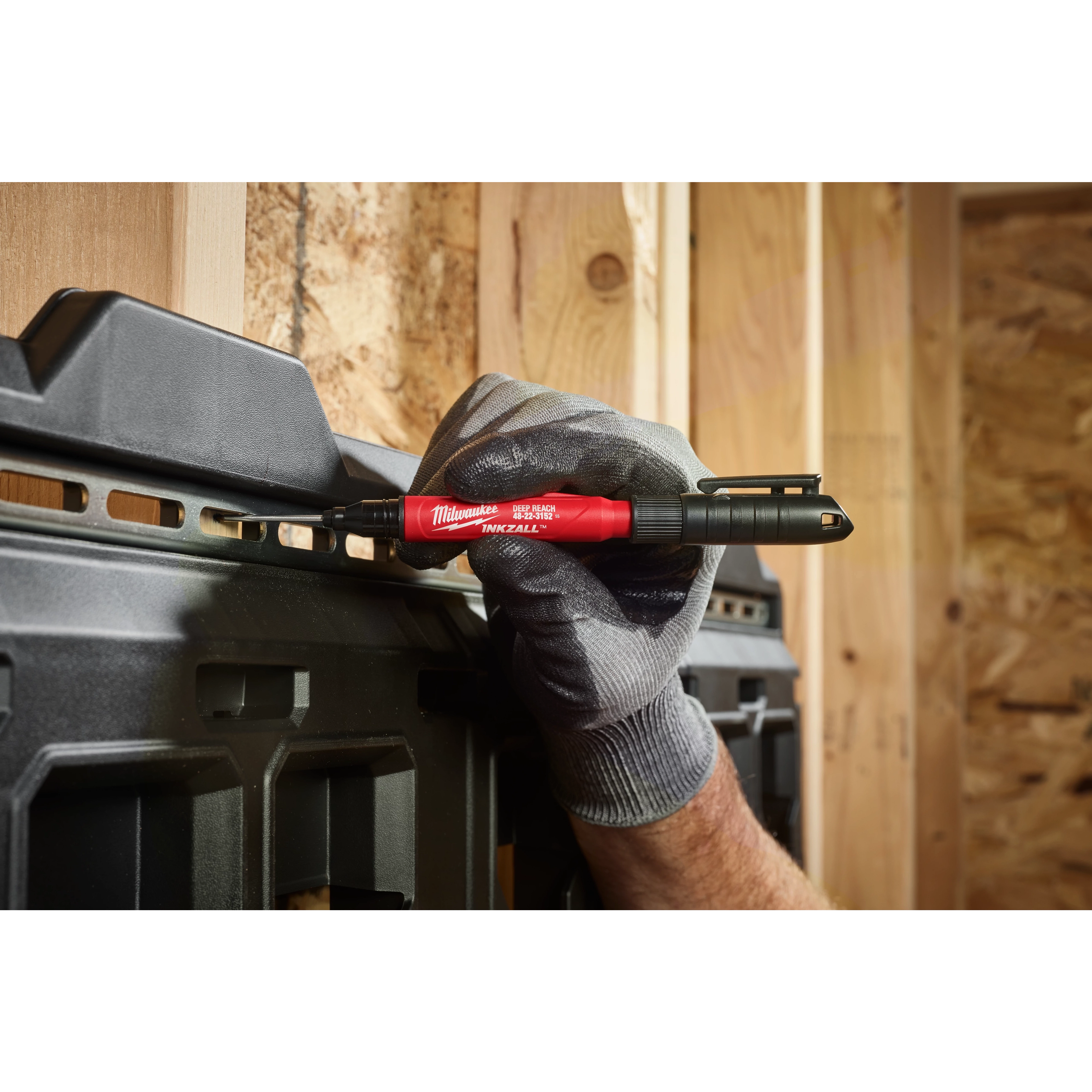 A gloved hand holding the INKZALL™ 2IN1 Deep Reach Ultra Fine Point Black Jobsite Marker is marking a dark plastic tool organizer mounted on a wooden wall. The marker has a red barrel with black details and branding displayed, highlighting its design suited for construction settings.