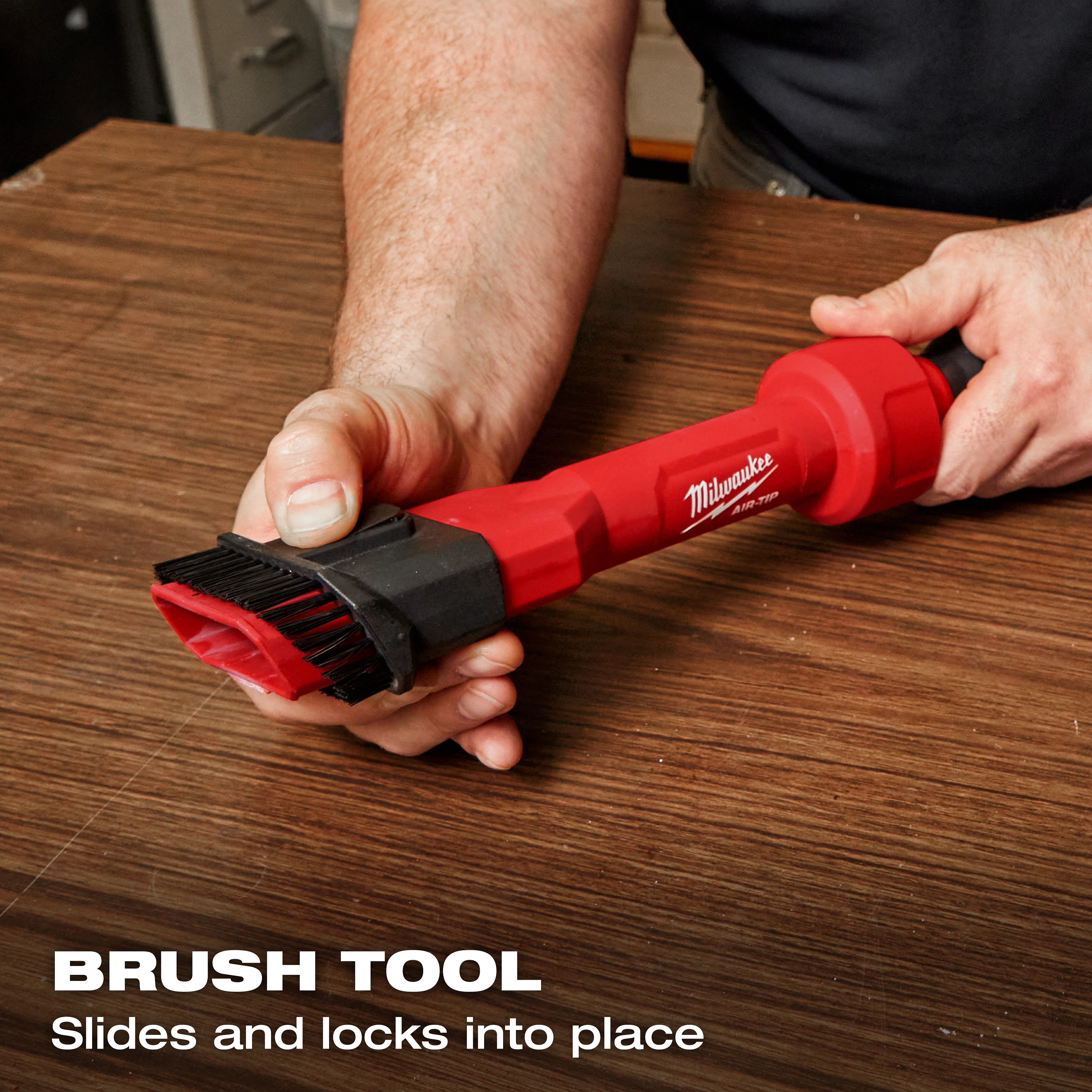 A person holding the red AIR-TIP™ 2-in-1 Utility Brush Tool on a wooden surface. The tool has a brush head on one end and is labeled with "Milwaukee" and "AIR-TIP™." Text overlay reads, “BRUSH TOOL: Slides and locks into place.”