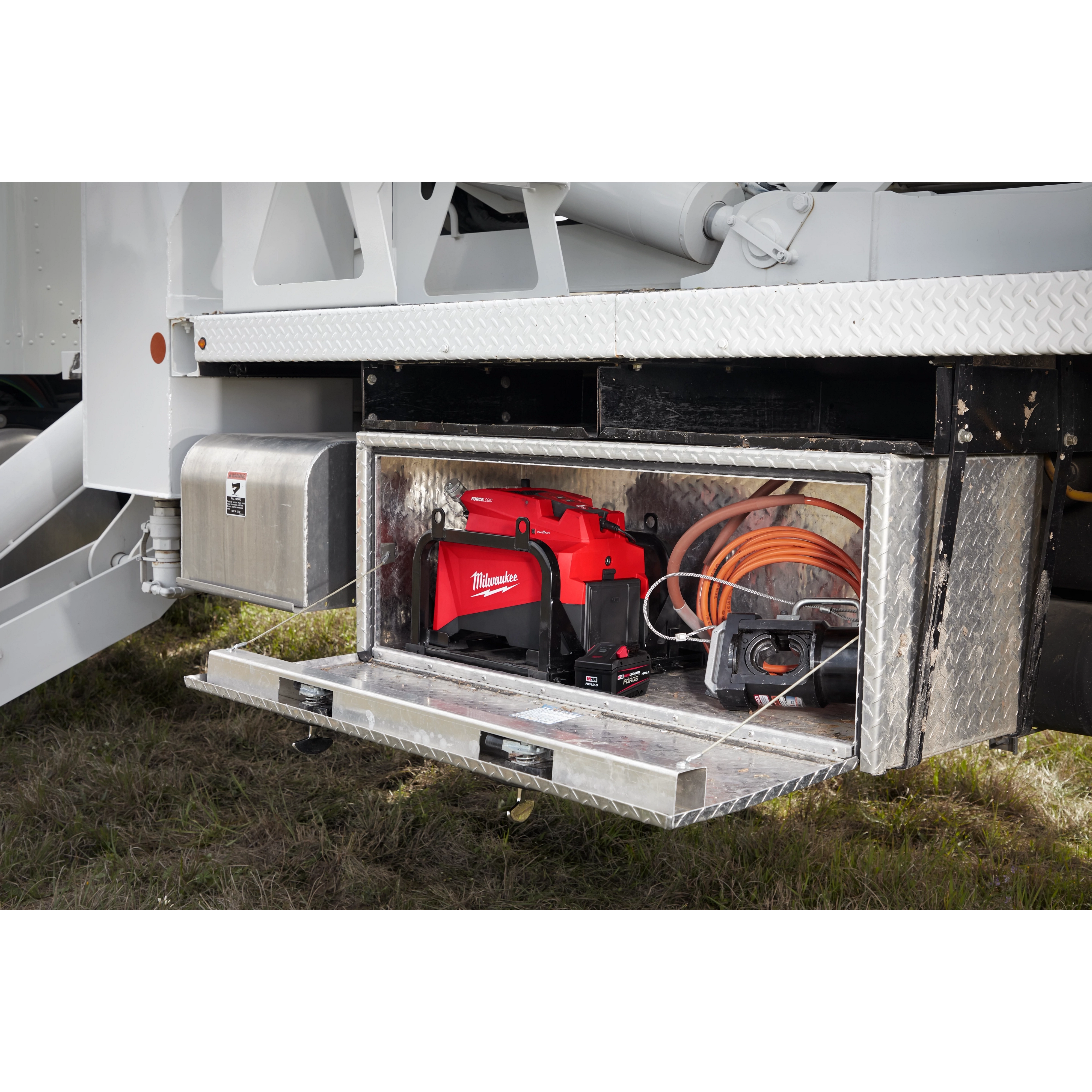 Open storage compartment on a vehicle containing an M18™ FORCE LOGIC™ 10,000 PSI Hydraulic Pump, a coiled orange hose, and additional electrical tools. The hydraulic pump is red and prominently positioned at the center of the compartment.