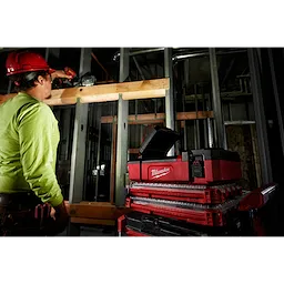 Worker using a drill at a construction site, with M12 PACKOUT Flood Light w/ USB Charging on a stack of red and black toolboxes nearby.