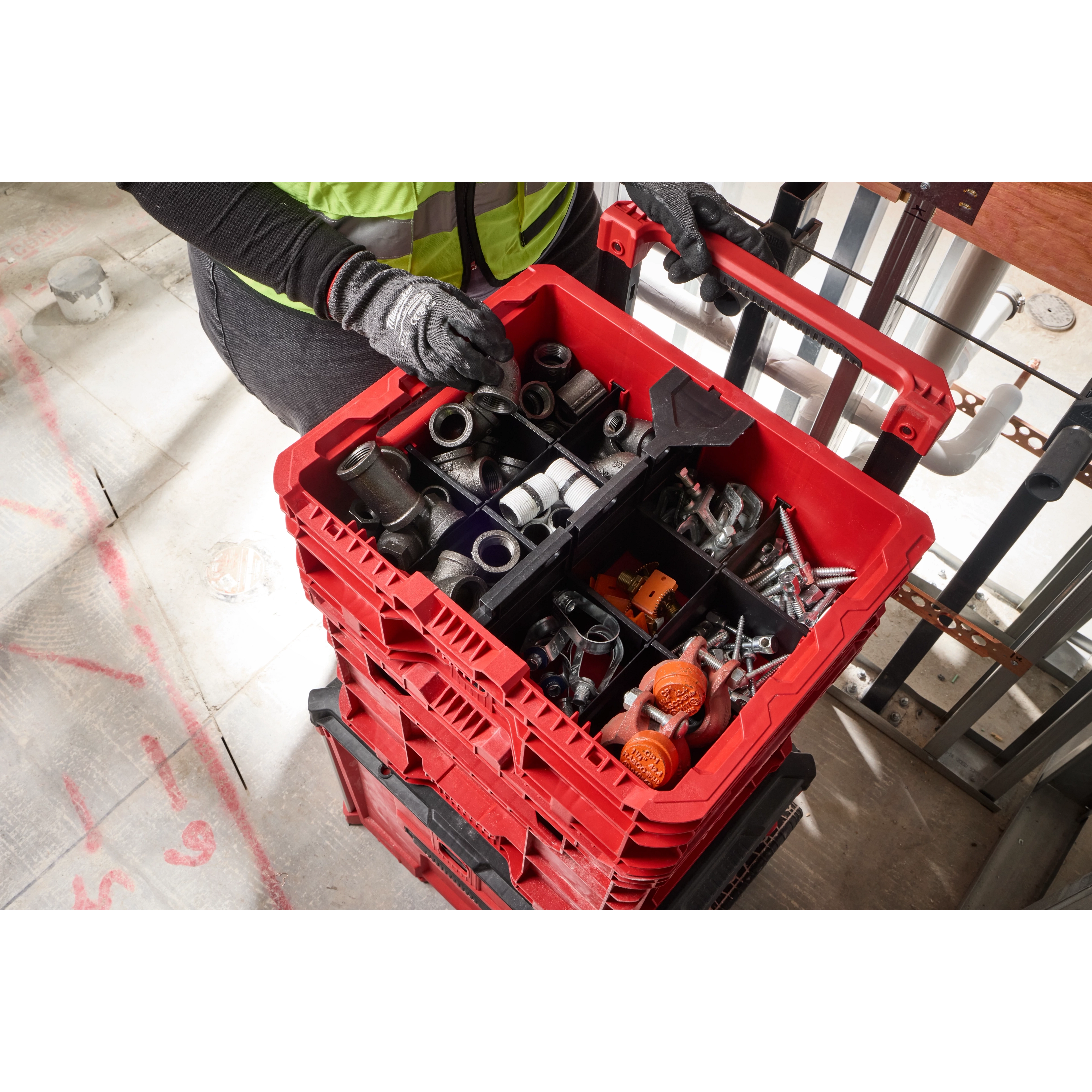 Person wearing gloves holding a red PACKOUT Low-Profile Crate filled with various tools and fittings, stacked on similar crates.