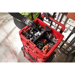 Person wearing gloves holding a red PACKOUT Low-Profile Crate filled with various tools and fittings, stacked on similar crates.