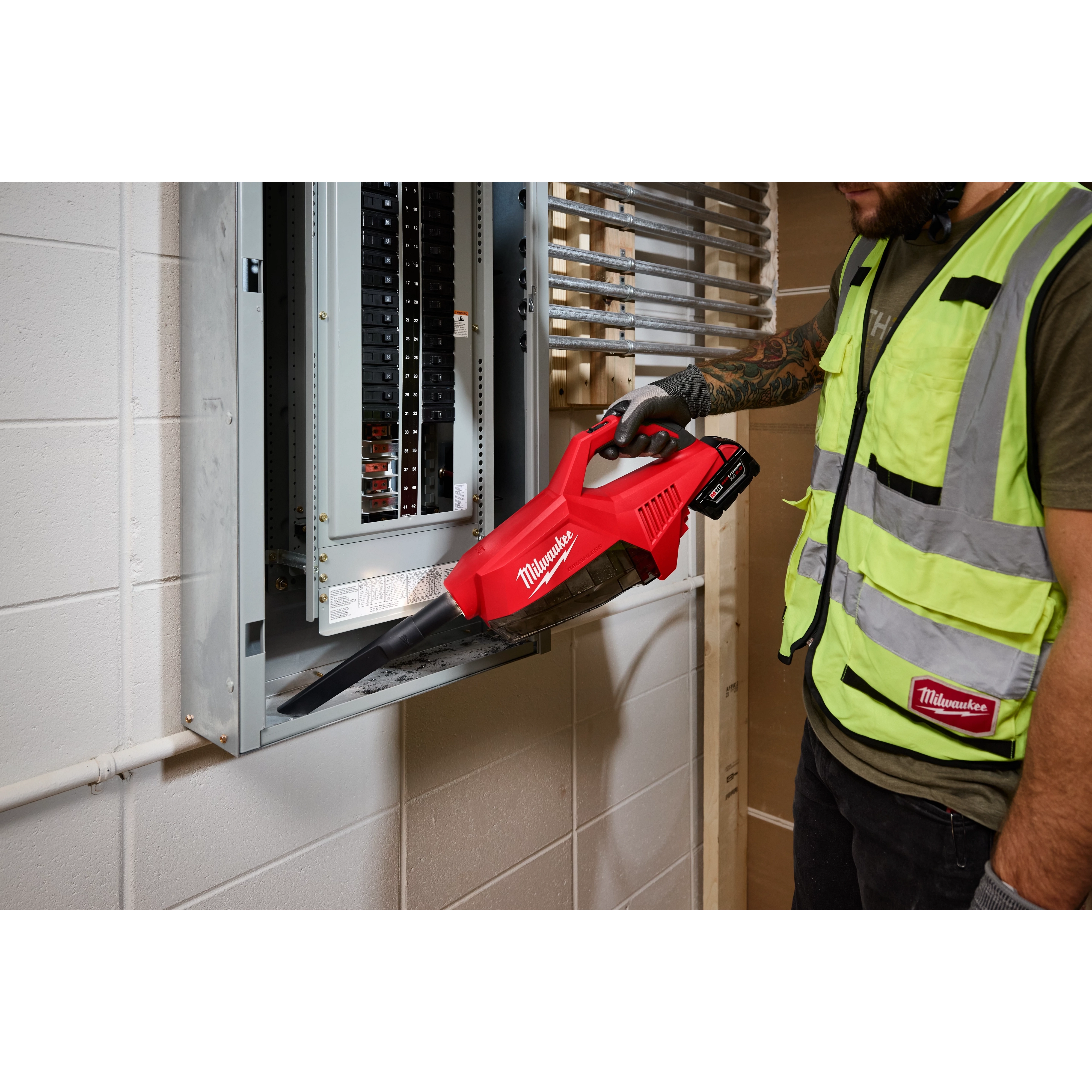 A worker uses the red M18 Brushless Handheld Vacuum to clean dust from an electrical panel. The vacuum is battery-operated and branded with the Milwaukee logo.