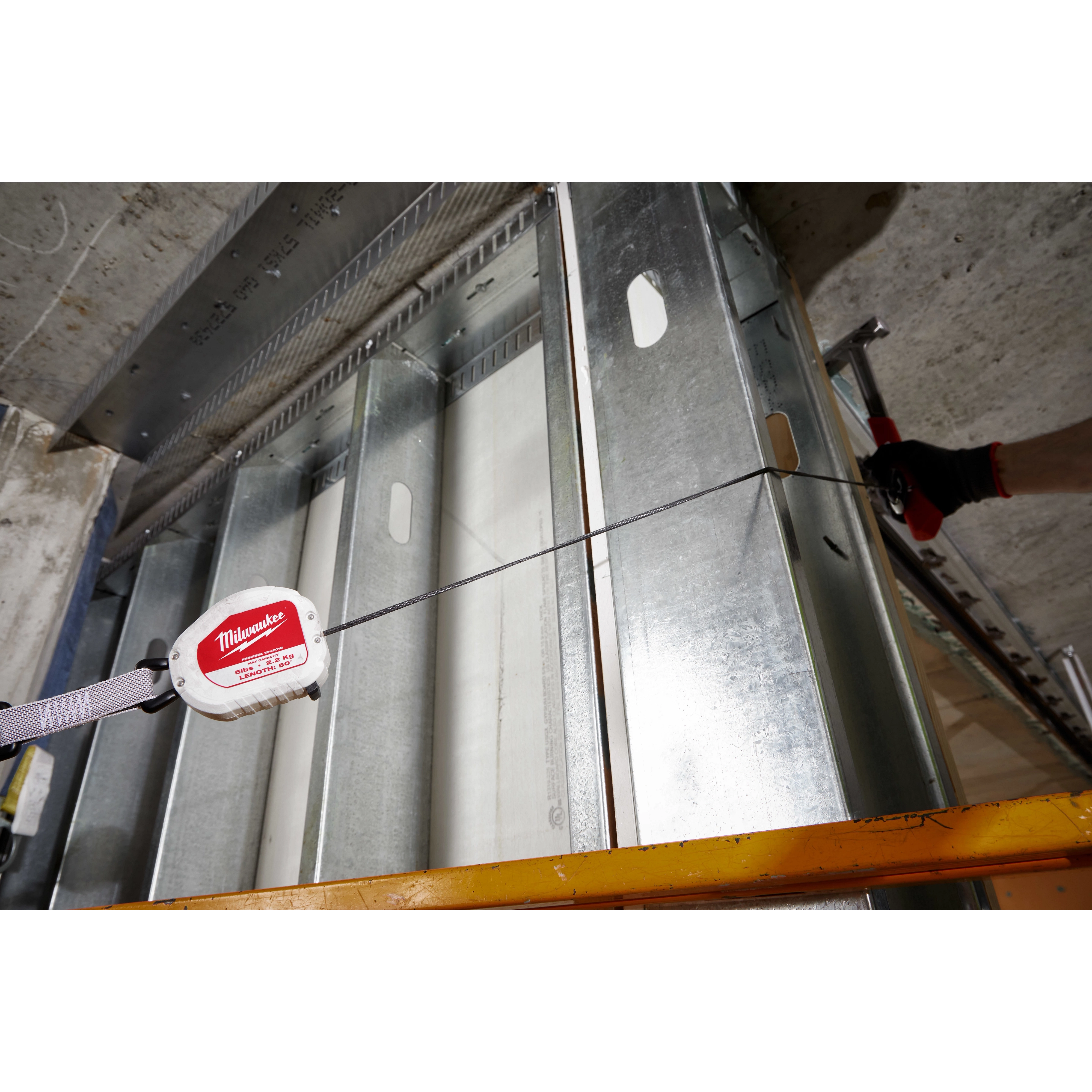 The image features a 5LB Quick-Connect Retractable Tool Lanyard attached to a steel frame construction. The lanyard is designed to secure tools and has a retractable cable extending from a white and red casing, providing convenience for tasks at elevated work sites.