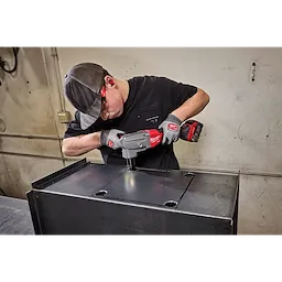 A man wearing safety gear uses an M18 FUEL™ 10 Gauge Nibbler to cut a metal sheet. The cordless tool is branded in red and is designed for precise cuts in heavy gauge materials.