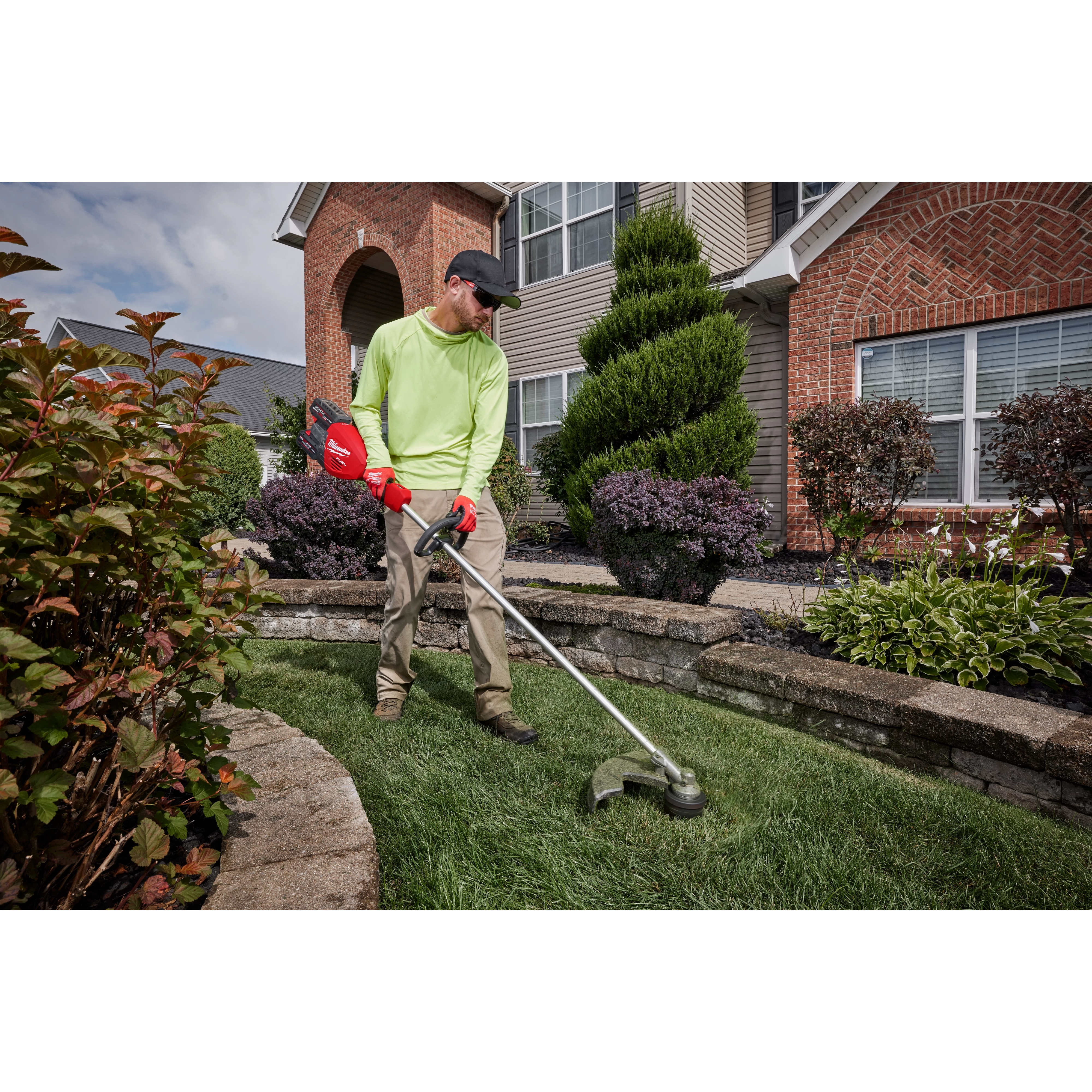 Person using a string trimmer to edge the lawn in front of a house surrounded by neatly landscaped plants.