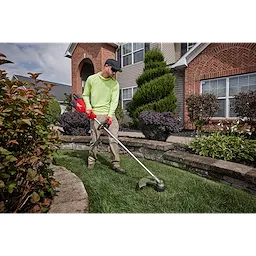 Person using a string trimmer to edge the lawn in front of a house surrounded by neatly landscaped plants.