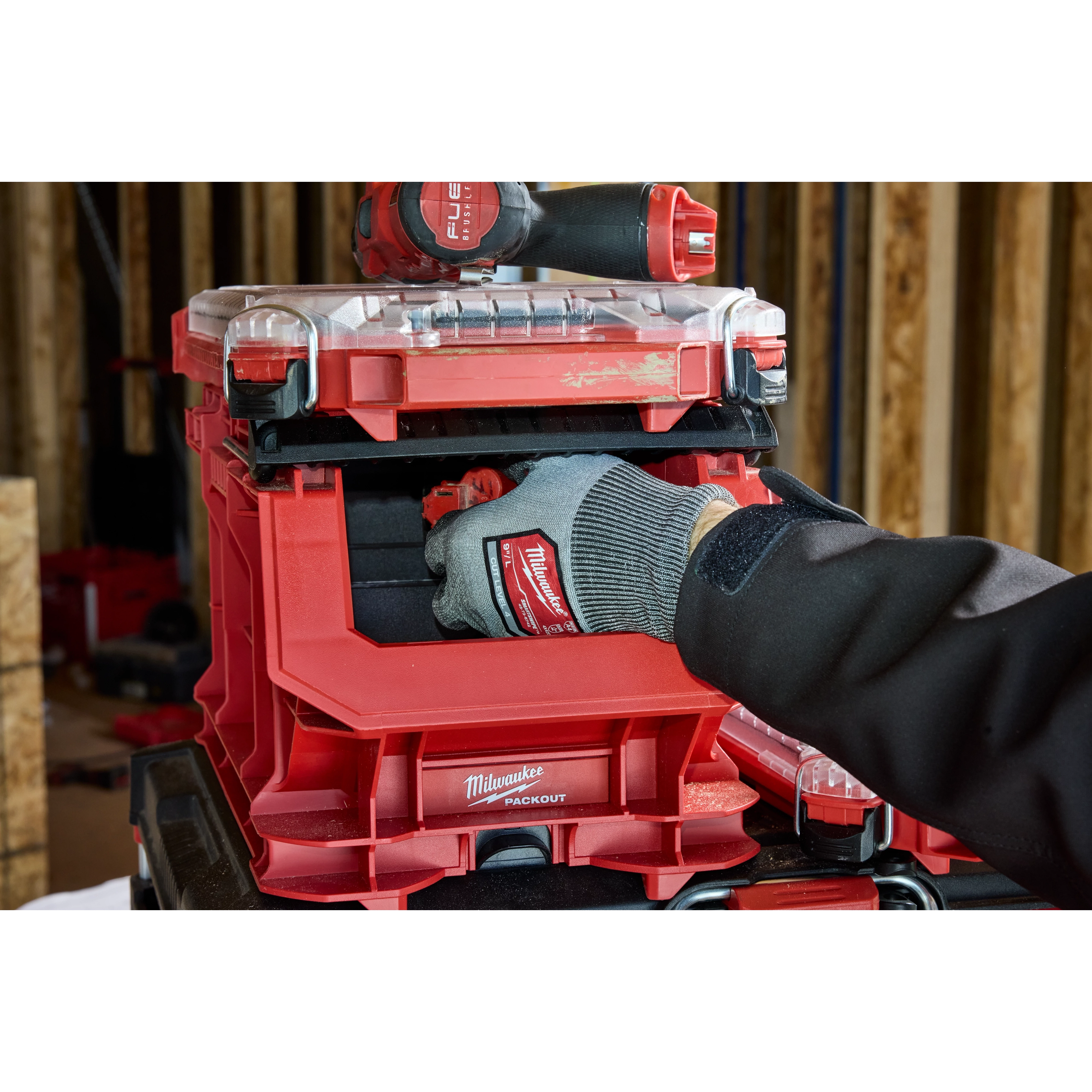 A person wearing a glove is organizing tools in a red PACKOUT Compact Crate with a clear lid, stacked in a workshop.