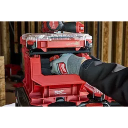 A person wearing a glove is organizing tools in a red PACKOUT Compact Crate with a clear lid, stacked in a workshop.