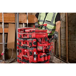 A person wearing work gloves and a high-visibility vest lifts a PACKOUT™ Tool Box Caddy Attachment in a workshop.