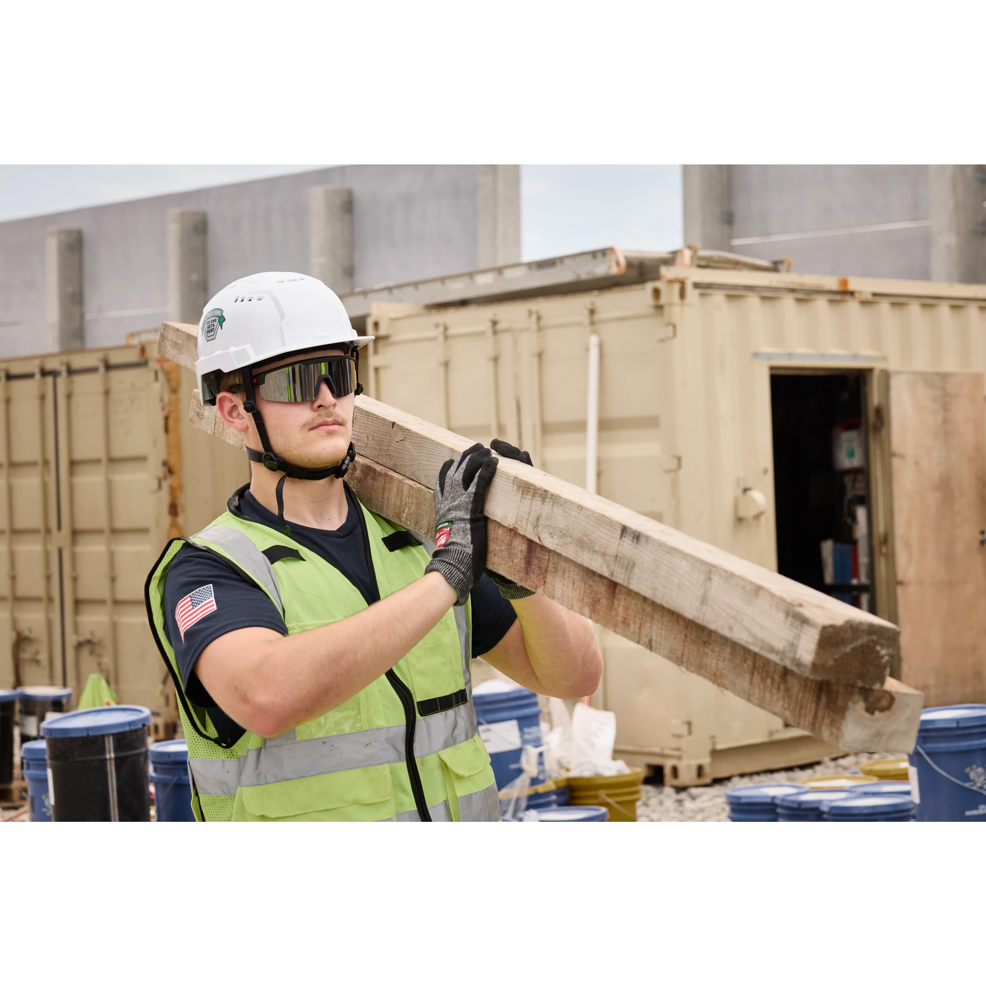 A construction worker in a safety vest and helmet carries a wooden plank on his shoulder. He wears Wrap Around Safety Glasses - Mirrored Anti-Scratch Lenses and black gloves at a busy construction site with containers and barrels in the background.
