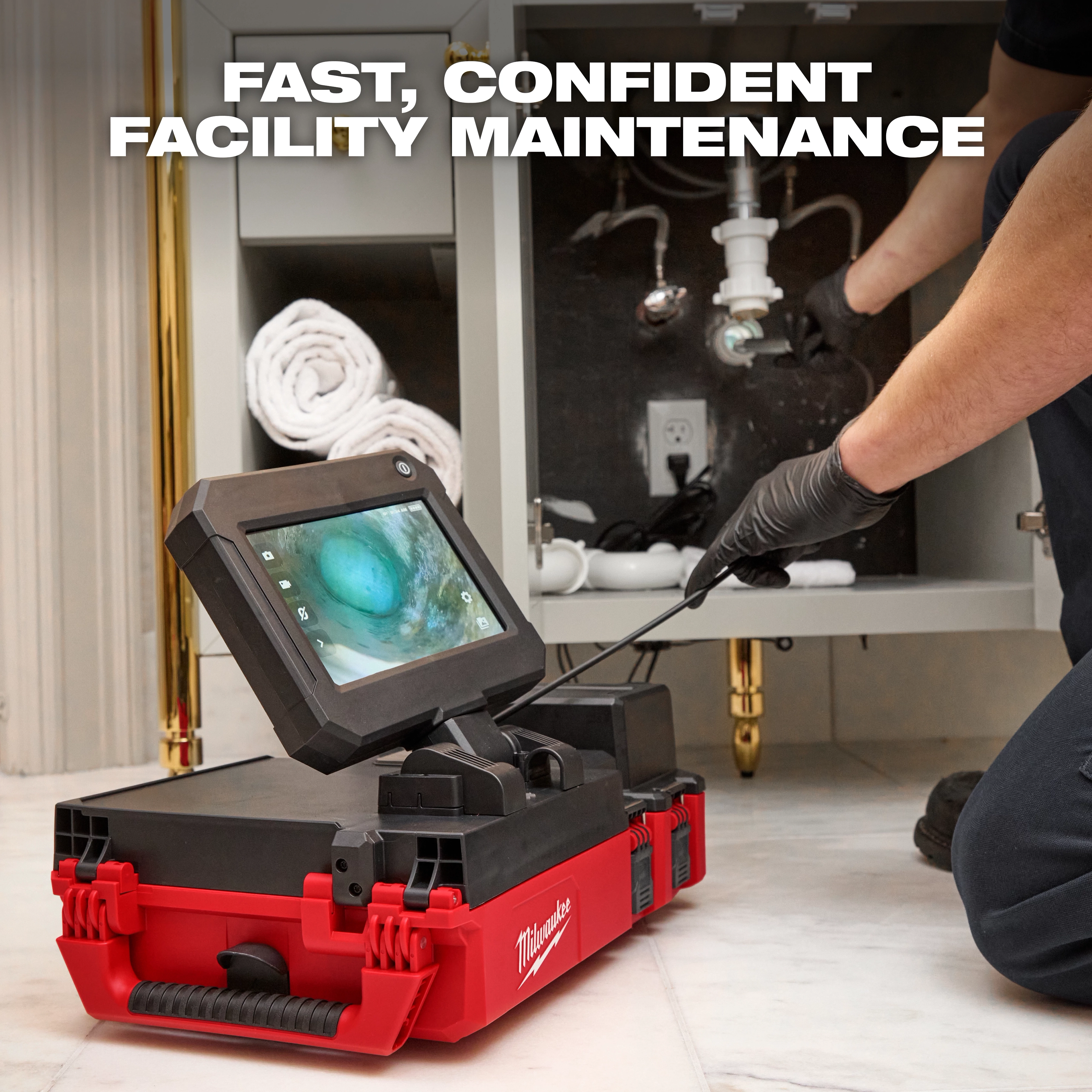 A person uses an M12™ 75' Drain Camera w/ PACKOUT™ Compatibility to inspect plumbing under a sink, with the camera display screen showing an image from inside the drain. The tool is placed on a red and black Milwaukee toolbox.