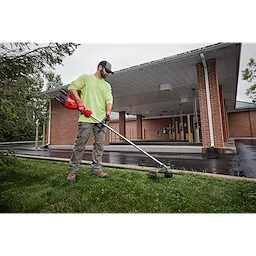 Person using a string trimmer to cut grass near a building with a covered driveway.