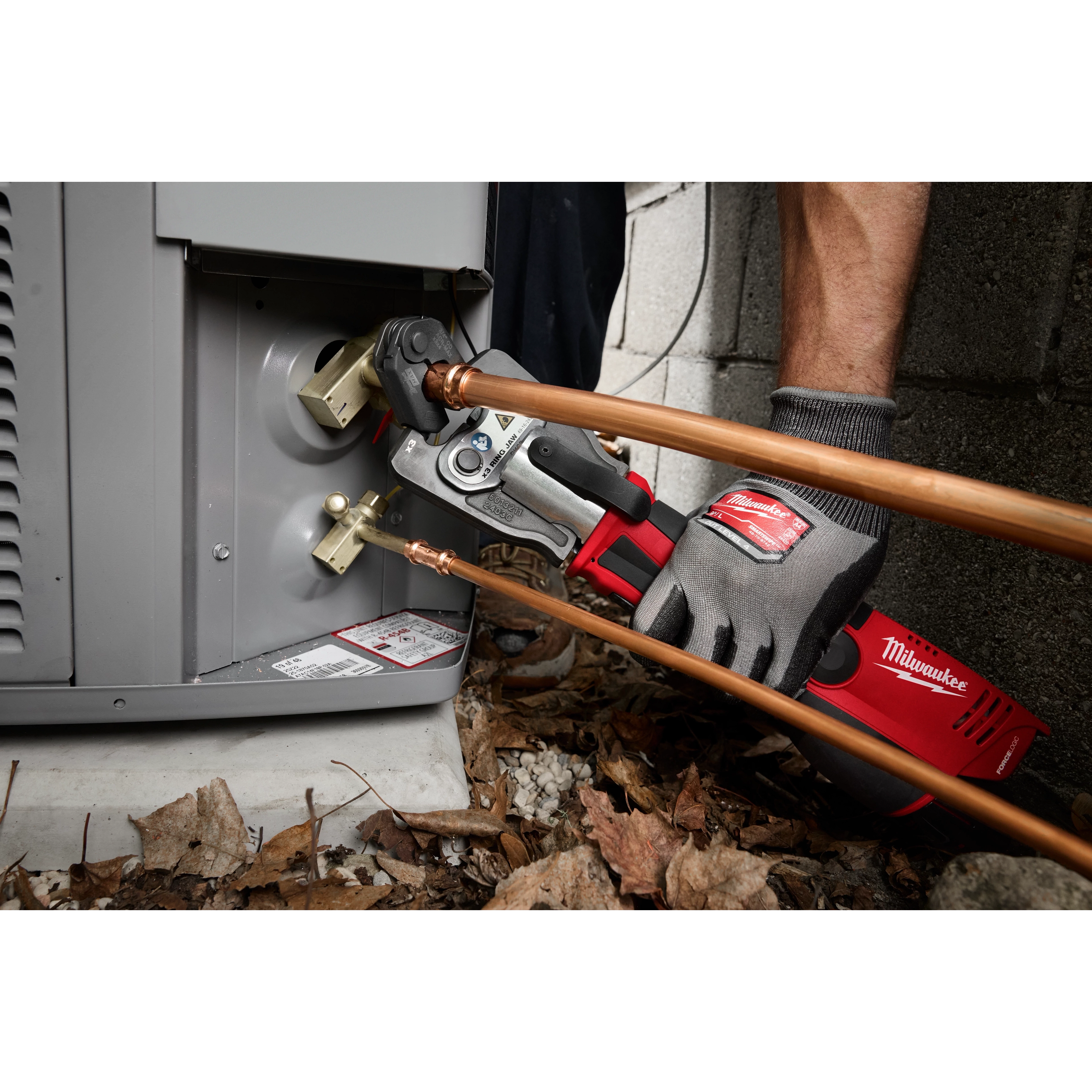 A technician uses a 3/4" ZoomLock MAX® & >B< MaxiPro® Pivoting Press Ring to connect copper pipes to an HVAC unit. The tool is connected to a red and black Milwaukee press machine.