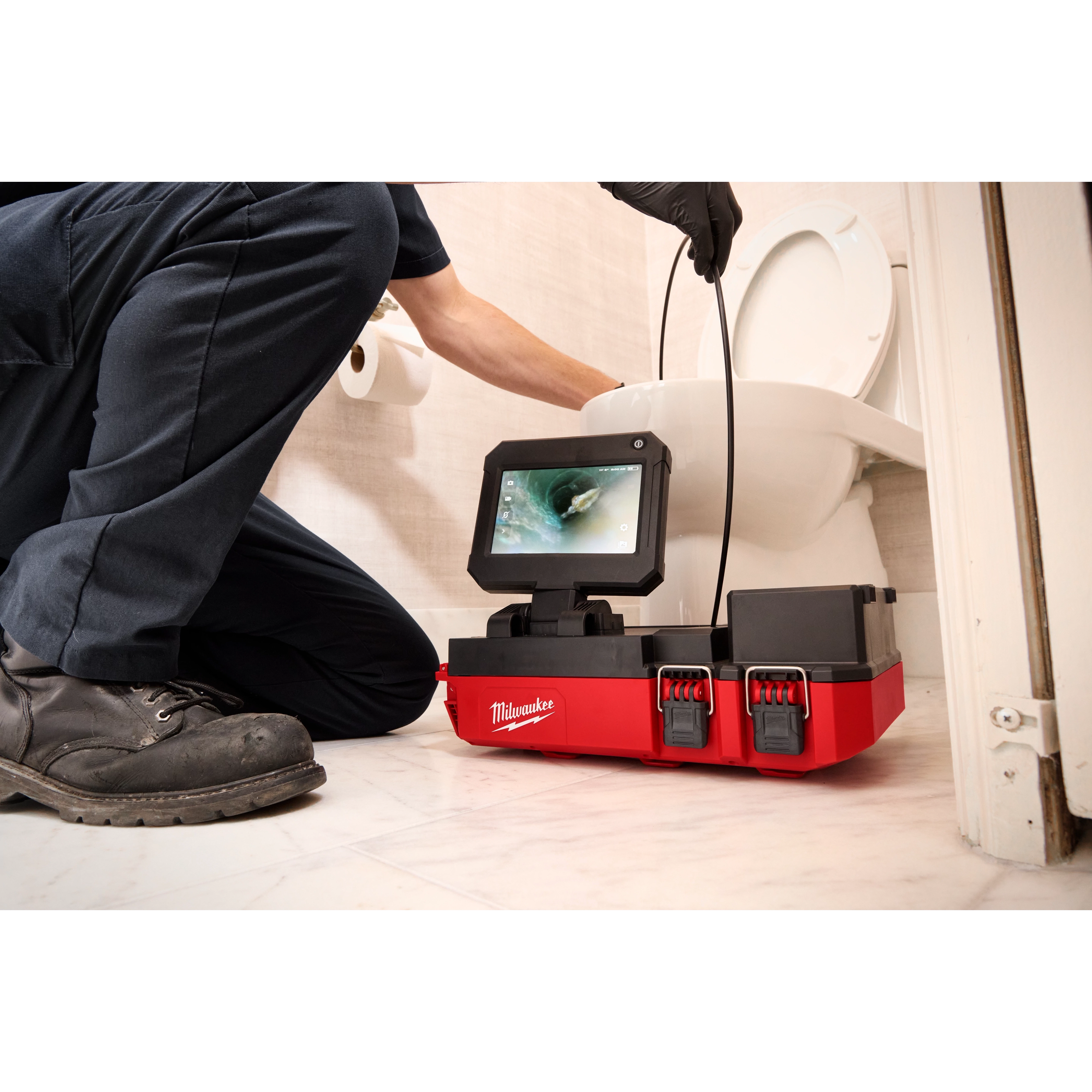 A person is using an M12™ 75' Drain Camera with PACKOUT™ Compatibility to inspect a toilet drain. The device, branded with "Milwaukee," features a screen displaying the camera feed and is housed in a red and black unit positioned on a bathroom floor.