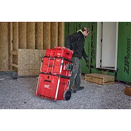 Person moving a stack of red PACKOUT Low-Profile Crate and other storage units on a wheeled cart in a construction setting.