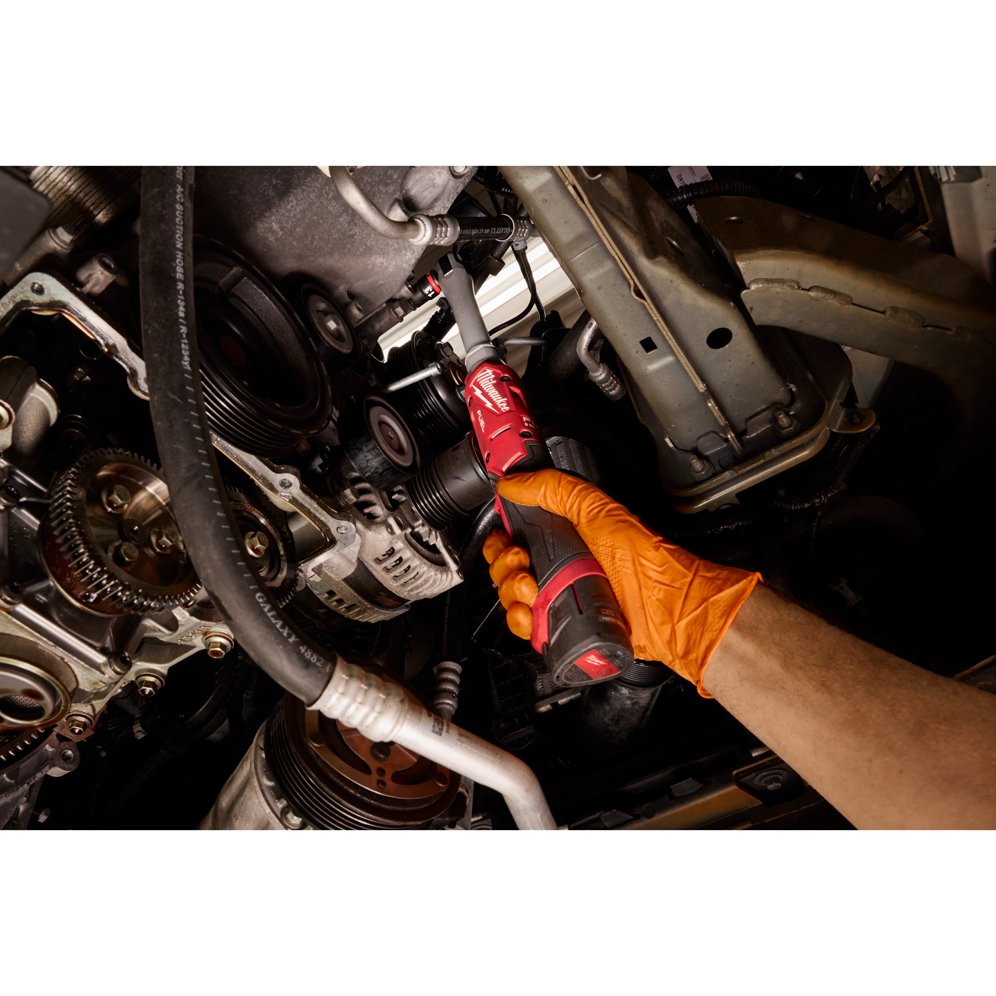 A person uses the M12 FUEL 3/8" Extended Reach Ratchet to work on a complex mechanical structure, possibly within an engine bay. The tool features a long, slender extension and ergonomic grip, designed for accessing hard-to-reach areas in tight spaces.