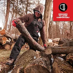 Person wearing FREEFLEX Insulated Bib Overalls, moving a log in a wooded area. Text reads "Designed to Layer Comfortably."