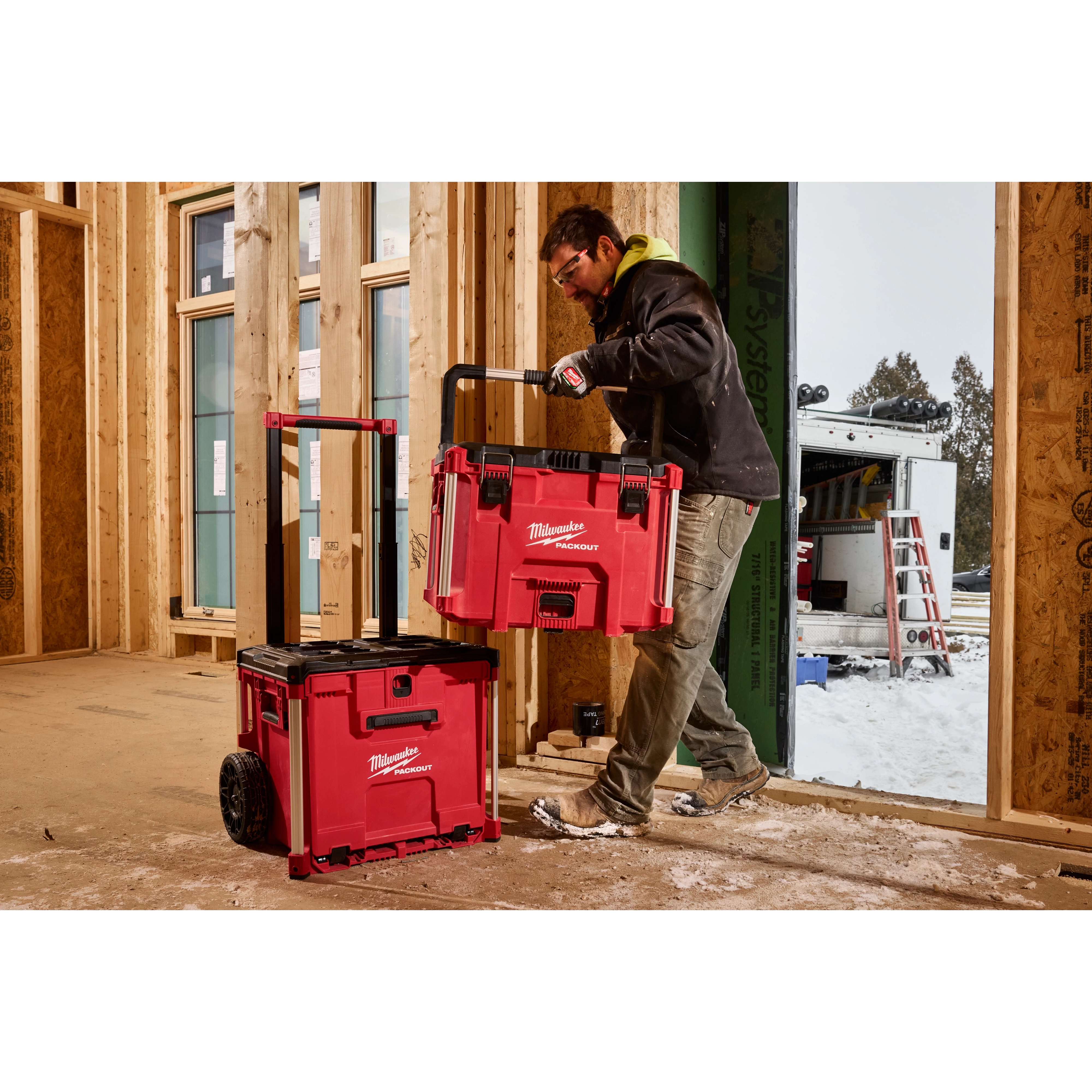 Person in a construction site handling a red PACKOUT XL Tool Box, stacking it on a wheeled base unit with a handle.