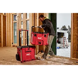 Person in a construction site handling a red PACKOUT XL Tool Box, stacking it on a wheeled base unit with a handle.