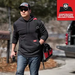A person wearing a dark gray hoodie and blue jeans carries a tool bag while wearing a GridIron Snapback Trucker Hat - Gray.